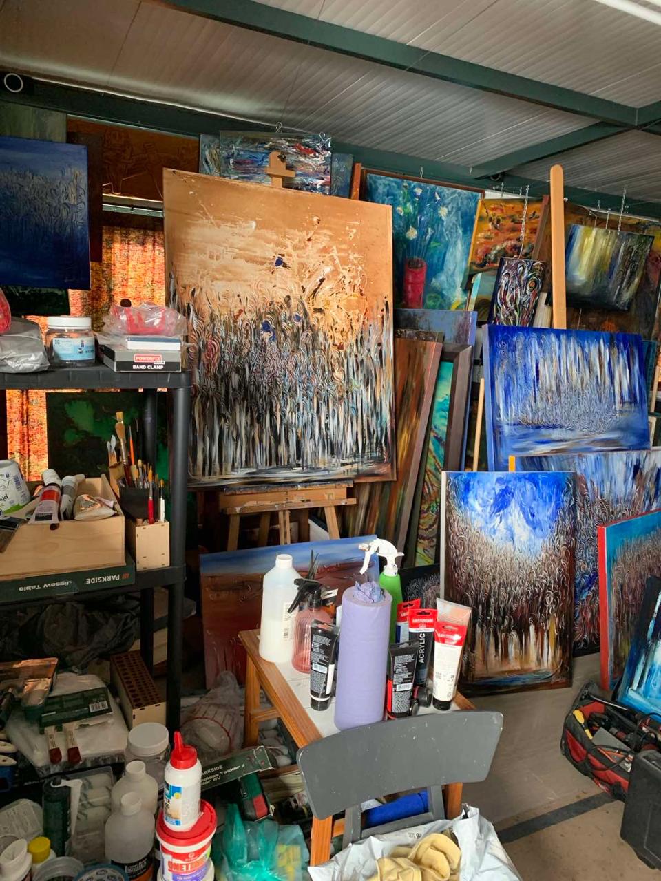 Workshop corner with abstract paintings on wooden easels, a small table holding brushes, paint tubes and bottles, boxes for a jigsaw table and a band clamp, and a tool bag on the floor beneath a paneled ceiling with green metal beams