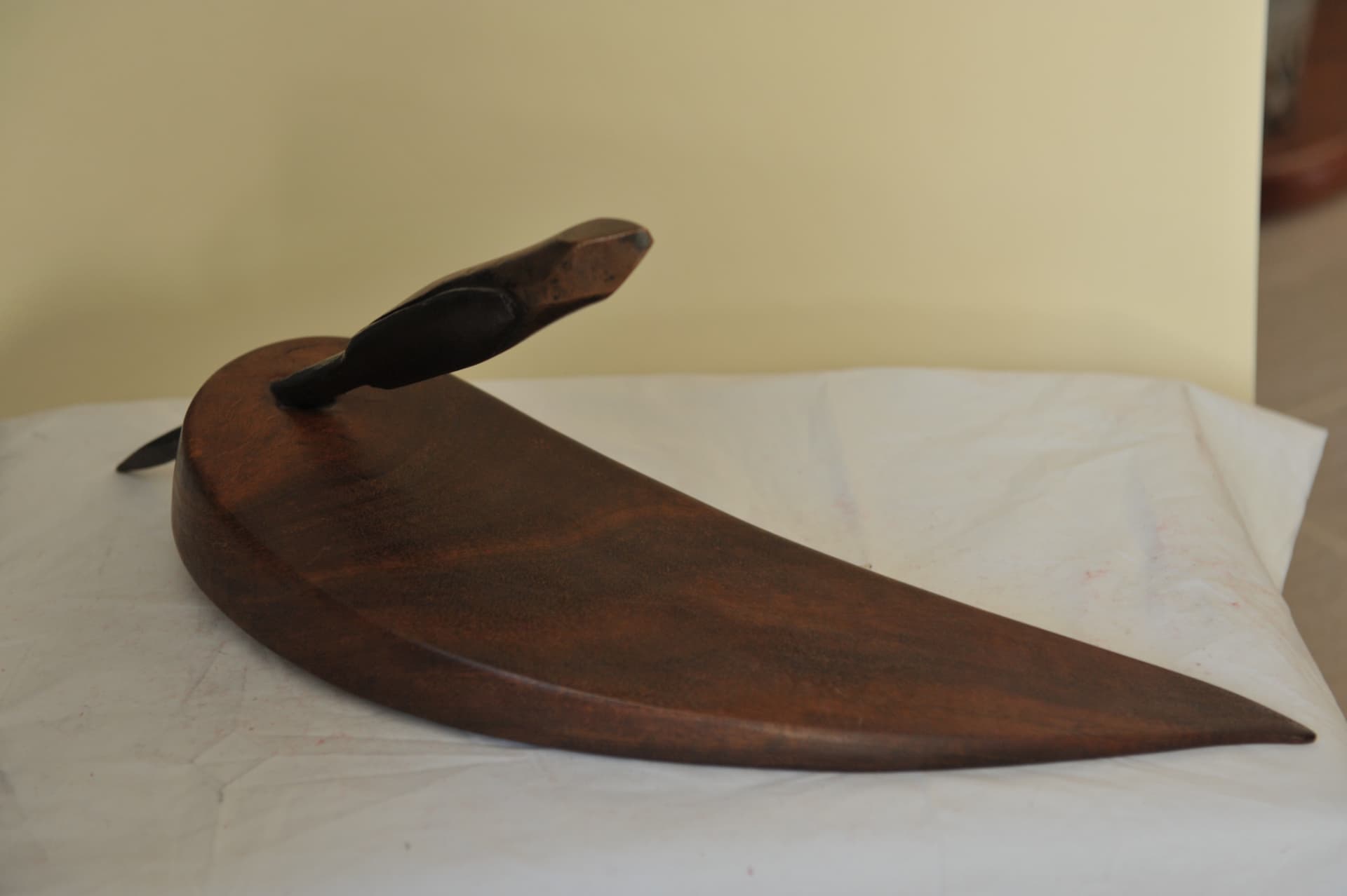 Curved wedge-shaped wood sculpture tapering to a long point, with a small angled projection at the thick end, resting on white cloth