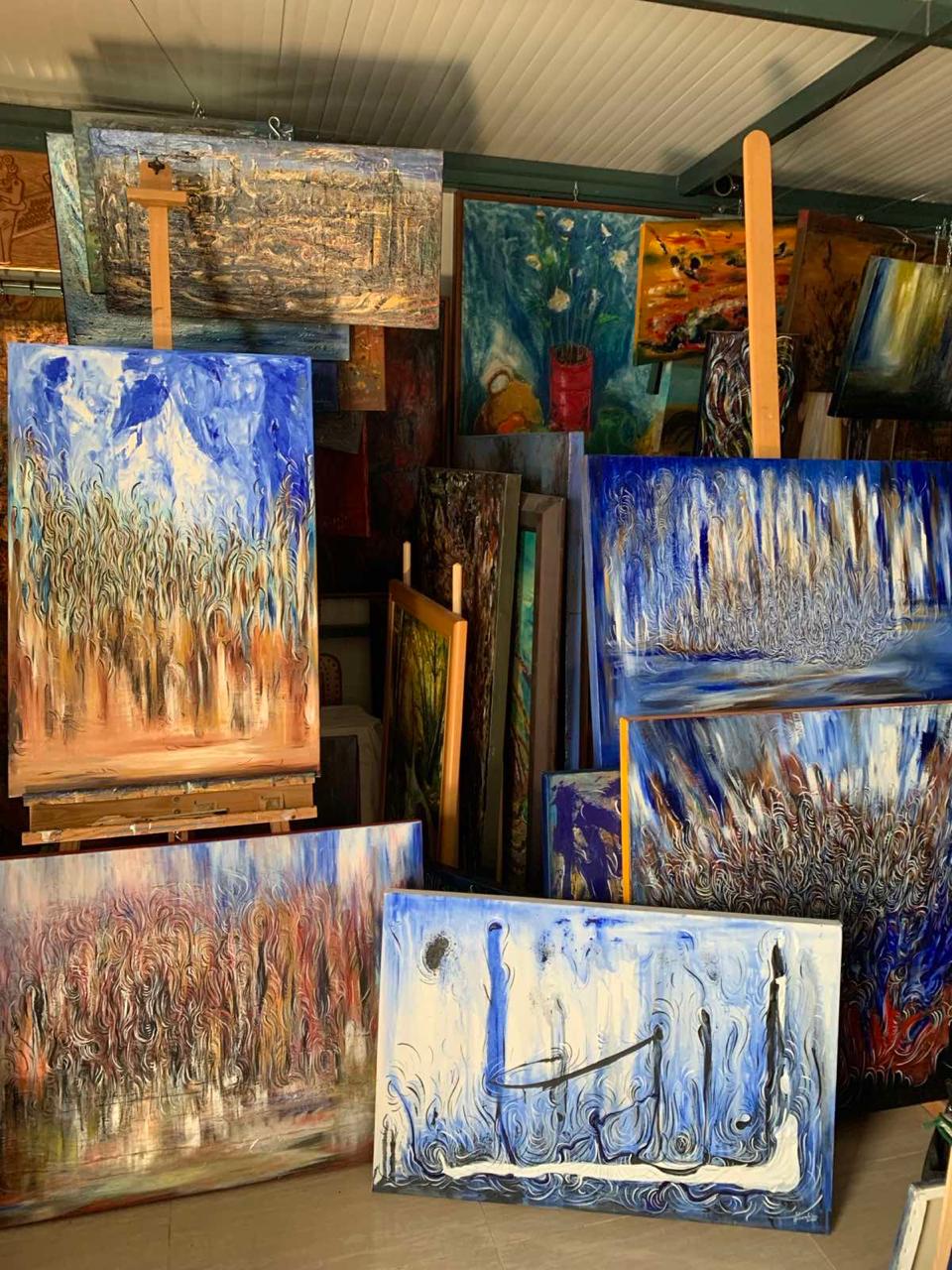 Studio interior filled with paintings on stretched canvases standing on tall wooden easels and stacked around the room, in deep blues, whites and earth tones, beneath a paneled ceiling with dark metal beams