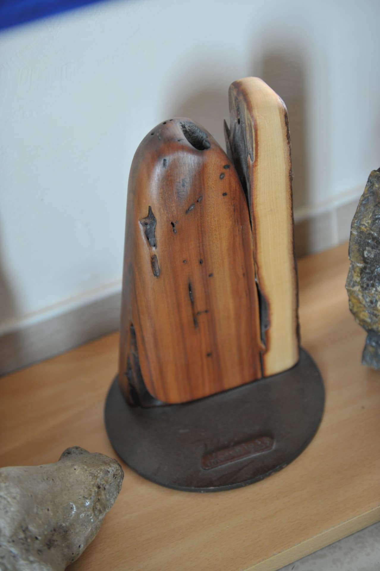 Upright two-part wood piece on a circular metal base, a domed block in front and a taller pale-faced section behind