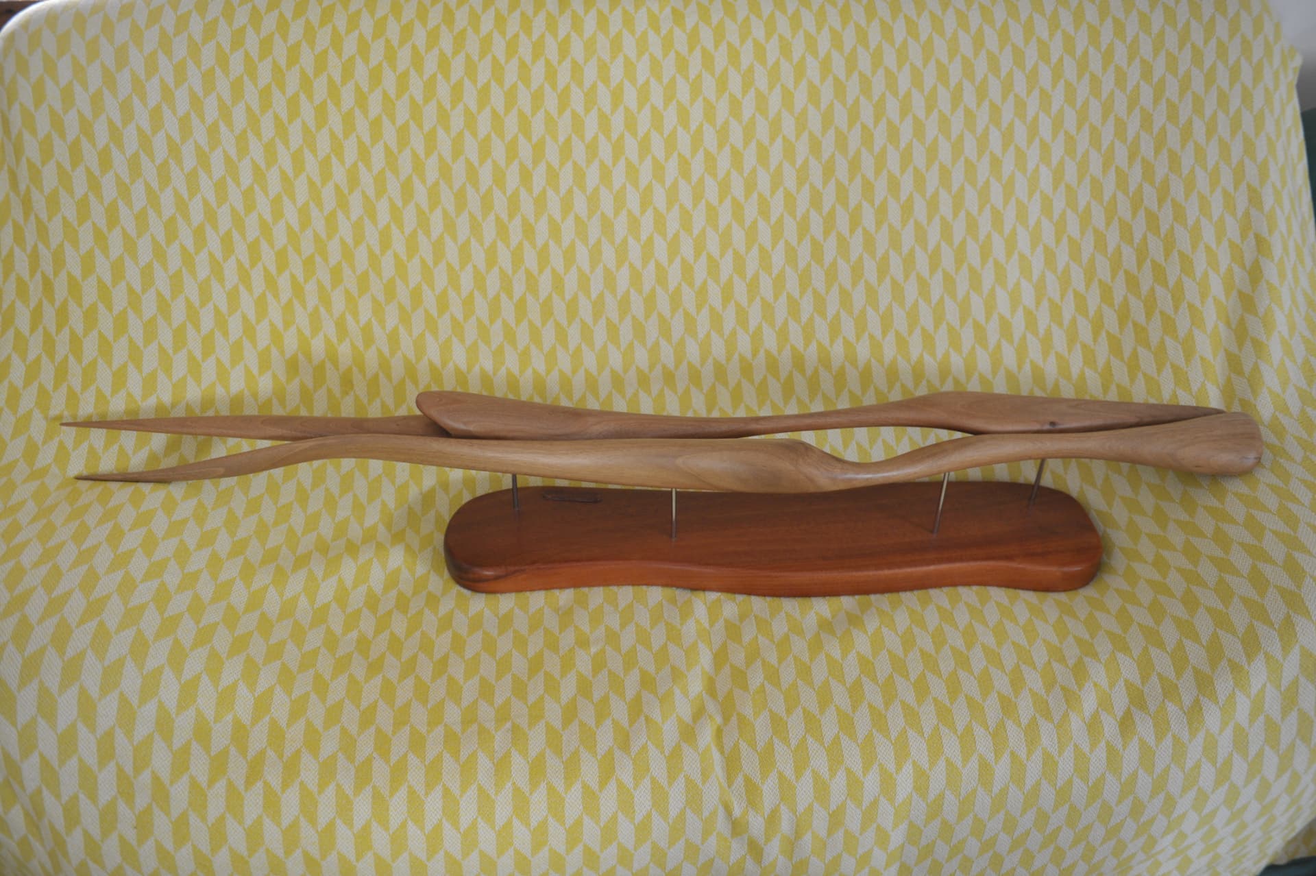 Two long tapering wood forms mounted on an elongated reddish base by thin metal rods, against a yellow and white patterned cloth