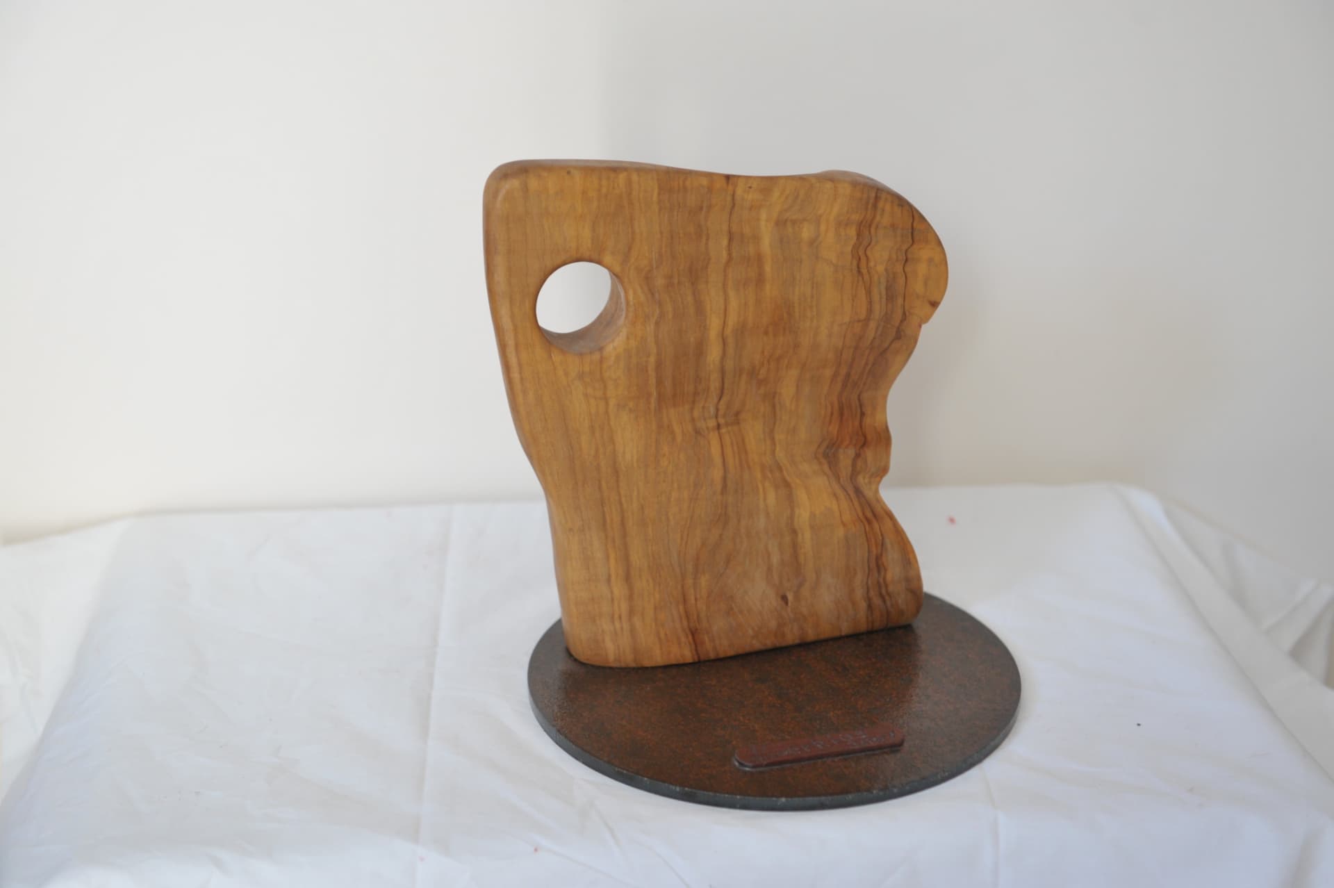 Flat vertical wood slab with a round hole near the upper left, mounted on a circular metal base against an off-white wall