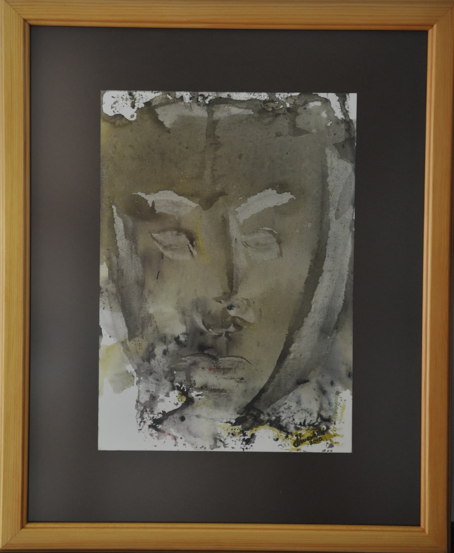 Face with closed eyes formed from pale paper highlights within charcoal grey wash and ochre accents, on paper mounted on a dark mat in a light wood frame