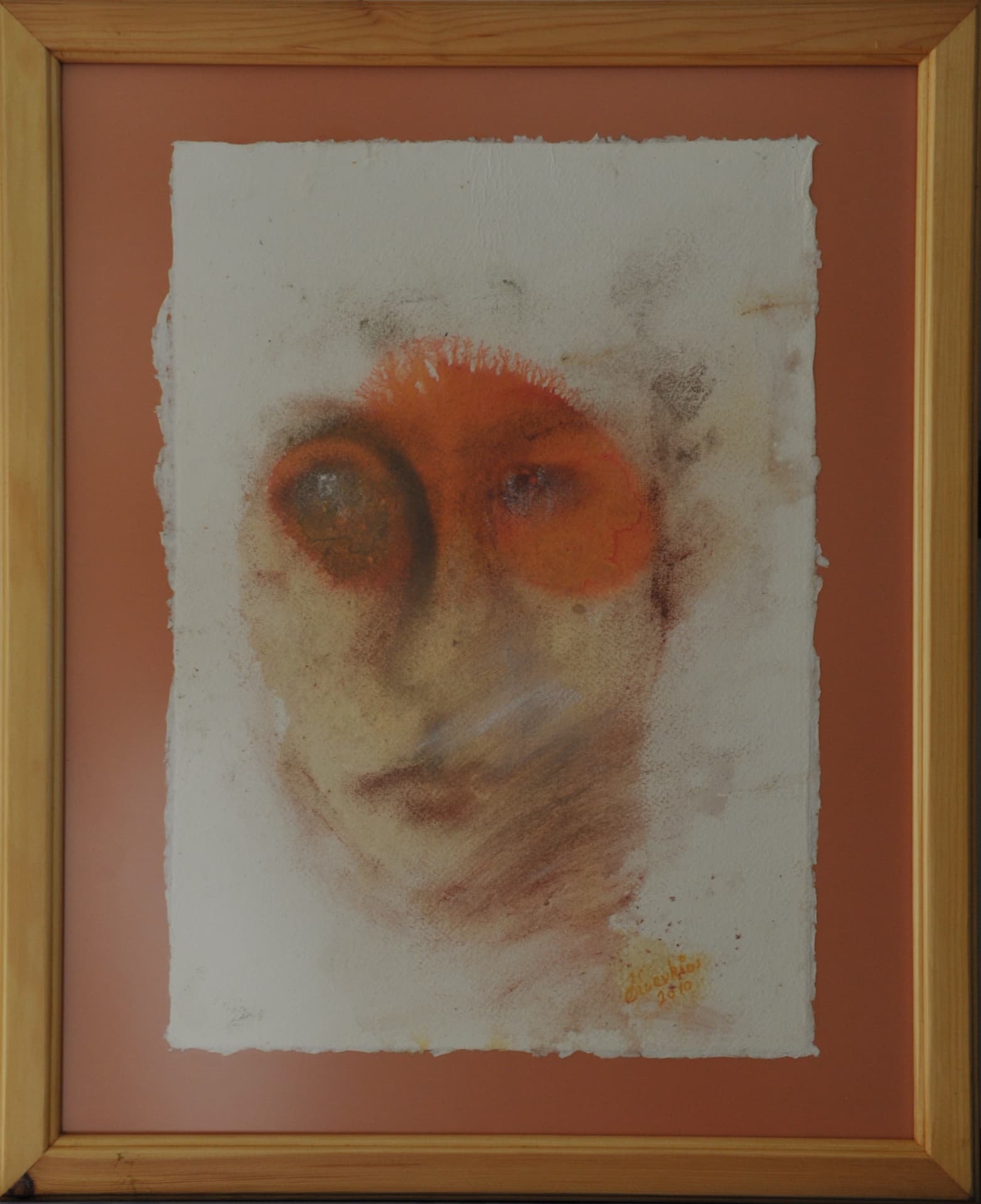 Face with circular markings around the eyes, in rubbed reddish-orange and brown pigment on deckle-edged paper mounted on a terracotta mat in a light wood frame