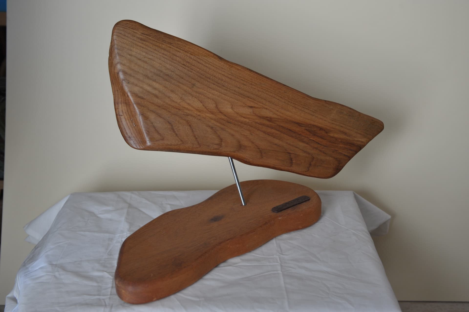 Two-part wood sculpture with an angled slab raised on a thin metal rod above a flat irregular base, against a cream wall and white cloth