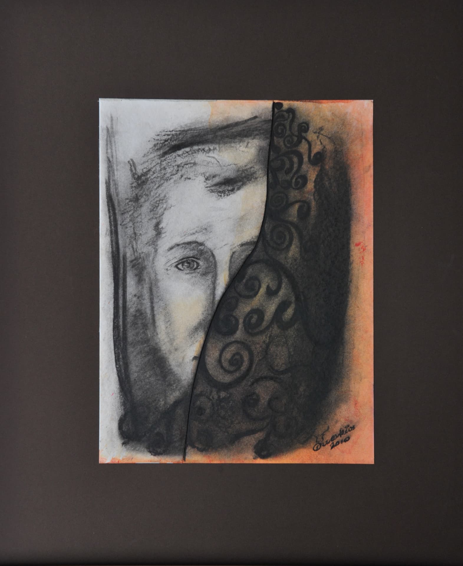 Two-part portrait: a pale face with one open eye in charcoal and grey wash beside dense black scroll patterns over orange and ochre wash, separated by a curved tear, on paper mounted on a dark brown mat