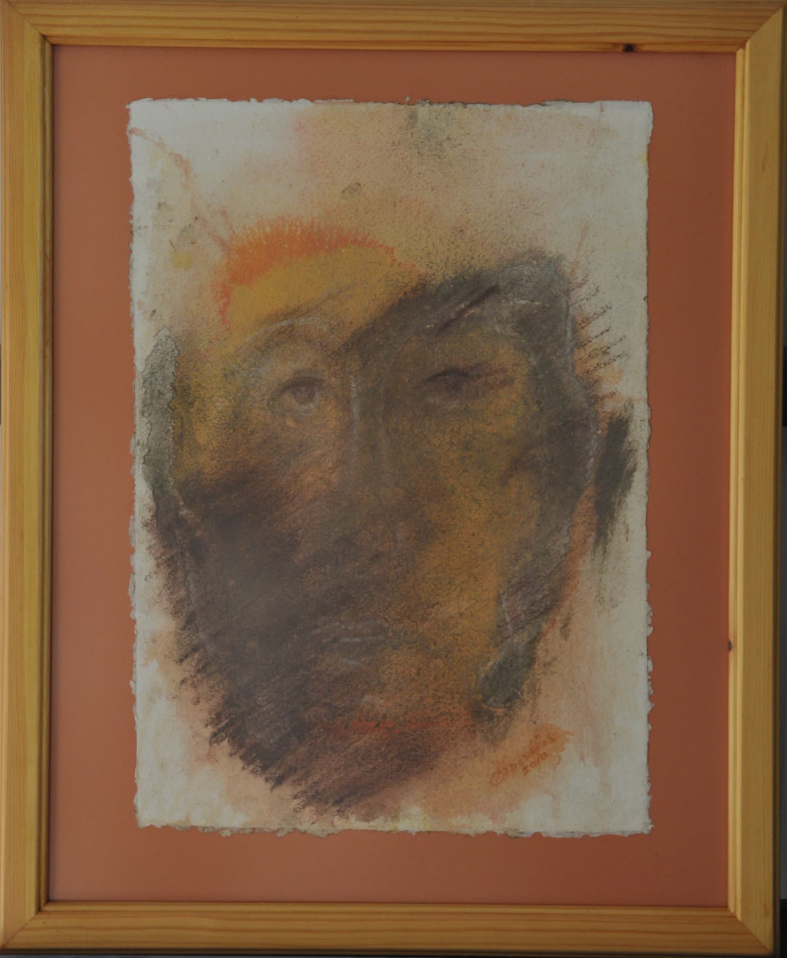 Frontal face in grained charcoal hatching beneath a halo of orange wash, on deckle-edged paper mounted on a terracotta mat in a light wood frame