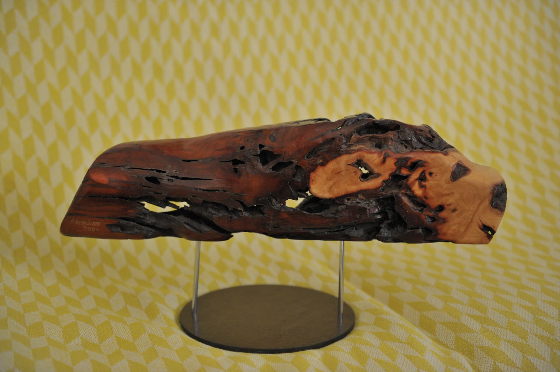 Horizontal dark wood sculpture with irregular openings on a circular metal stand
