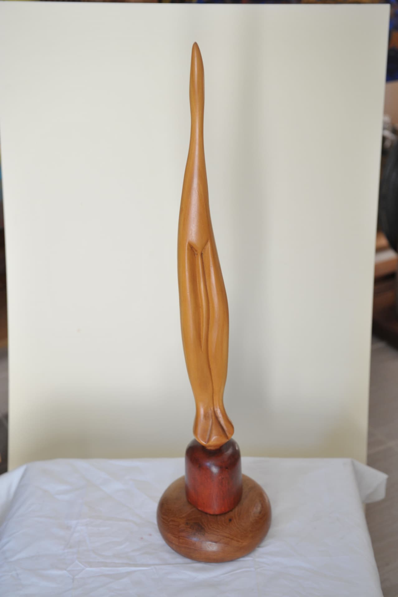 Tall slender tapering wood sculpture with a sharp tip and carved vertical folds, standing on a dark cylindrical riser and round base over white cloth