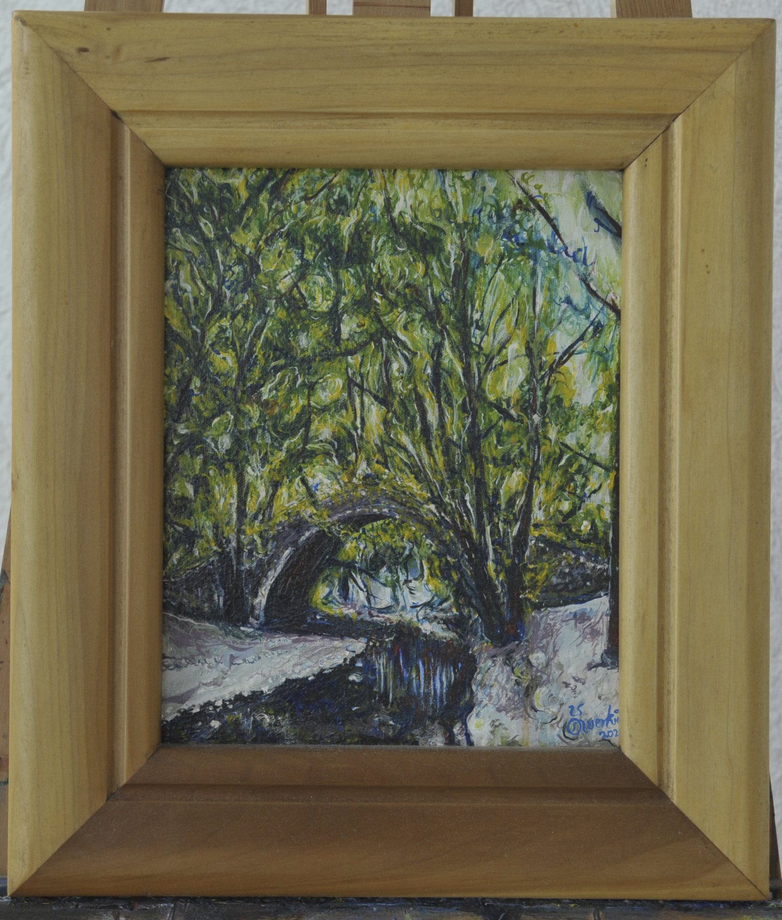 Painting of a stone arch bridge over water among green trees, in a wooden frame