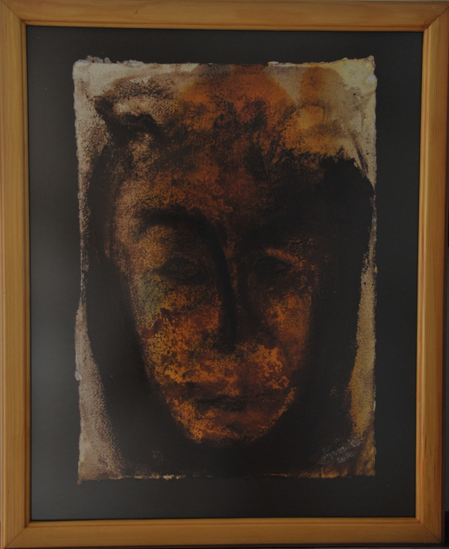Frontal face with closed eyes in granular dark rubbing over burnt orange wash, on deckle-edged paper mounted on a dark mat in a light wood frame