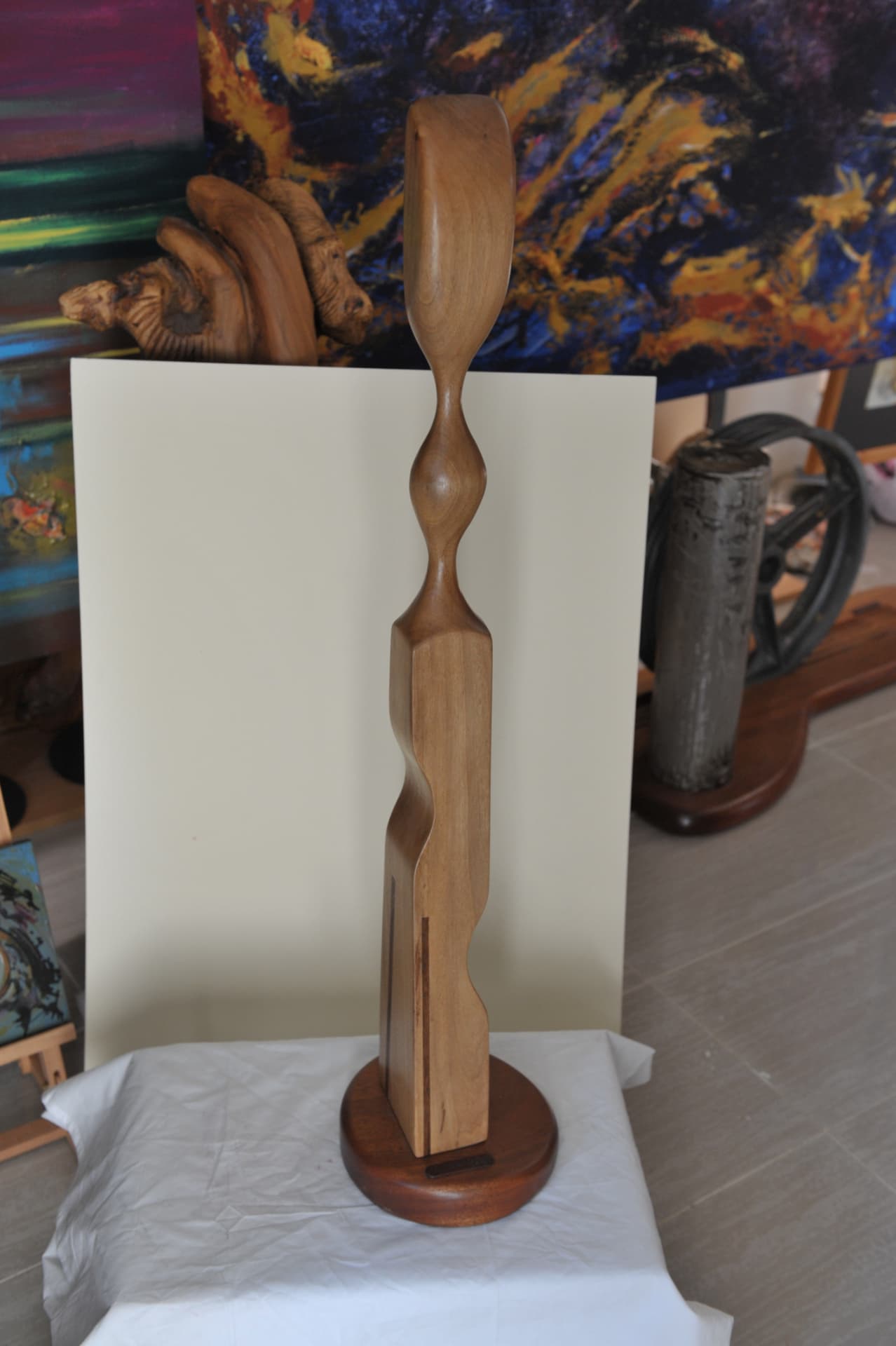 Totem-like wood sculpture with a pinched waist, a spherical nodule and an elongated rounded head, inlaid with dark stripes, on a thick circular base in a studio