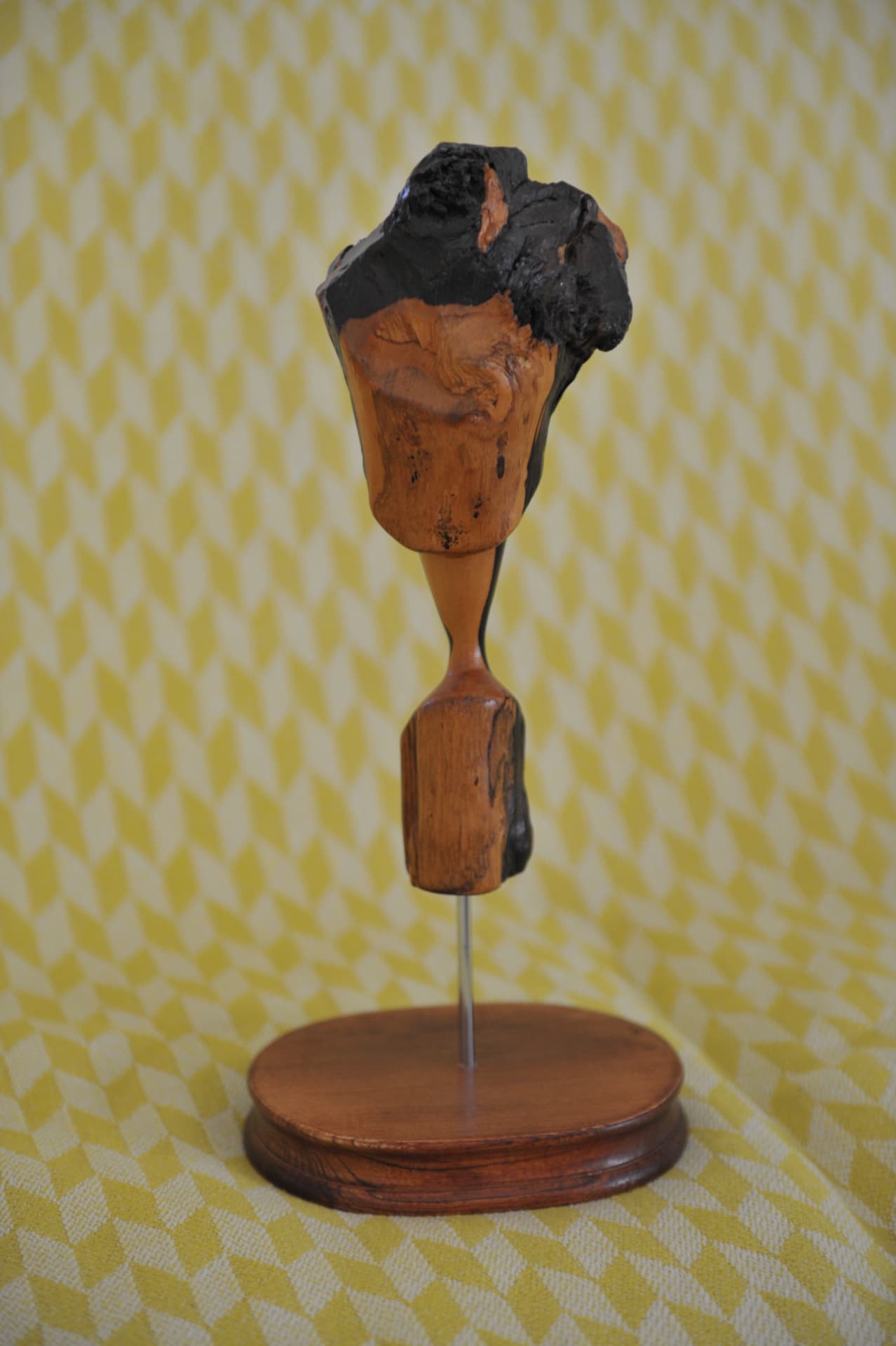 Upright wood sculpture with a bulbous blackened upper mass and a rectangular lower block joined by a carved waist, on a thin rod and oval reddish base against a yellow and white patterned cloth