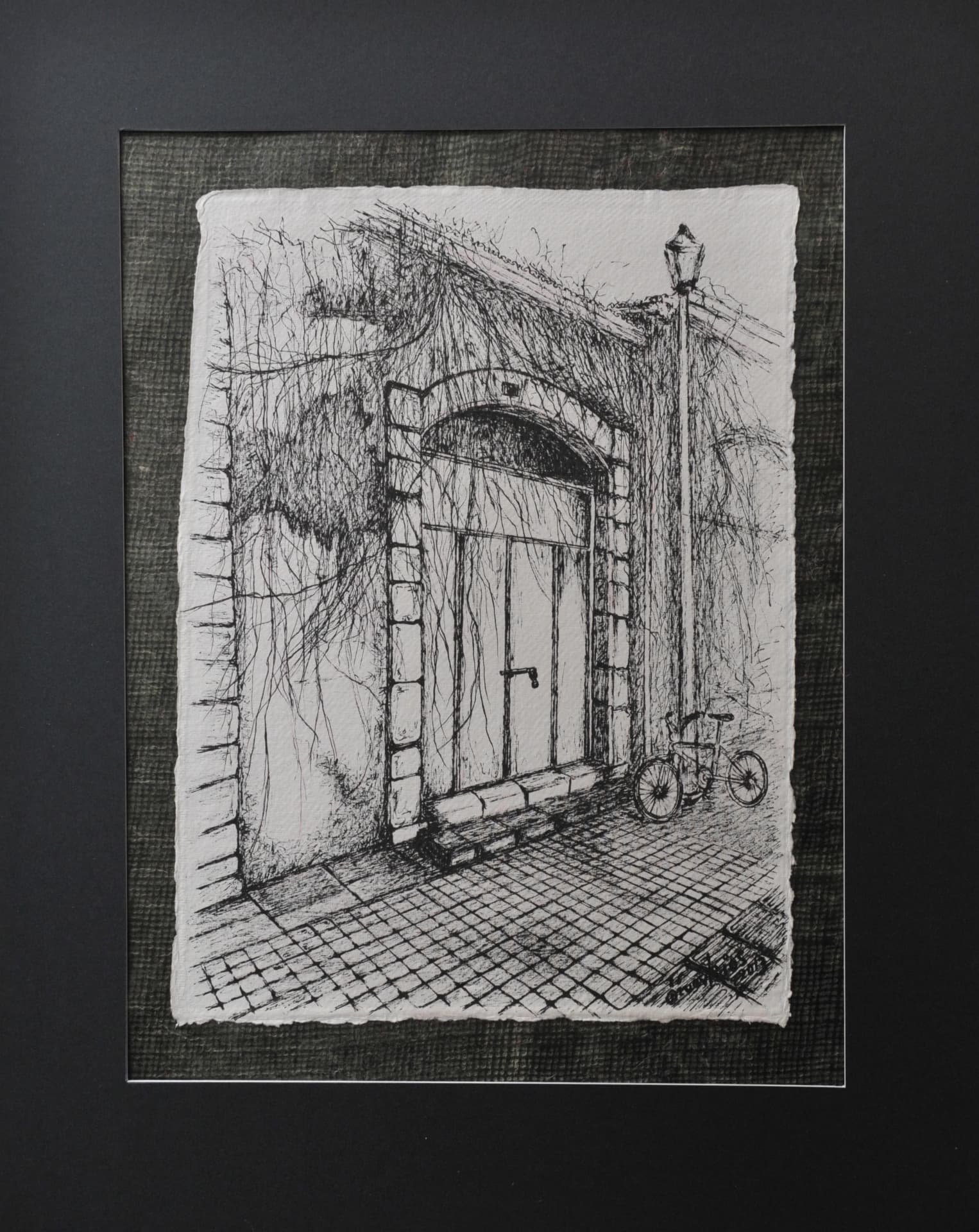 Ink drawing of a vine-covered stone archway door with a street lamp and a parked bicycle on a paved street, on paper mounted on a dark grey mat